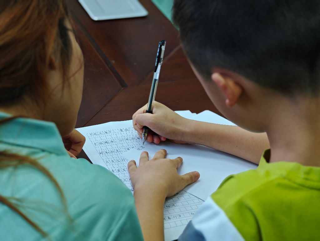 志愿者俯身陪伴学生书写练习,耐心指导汉字书写